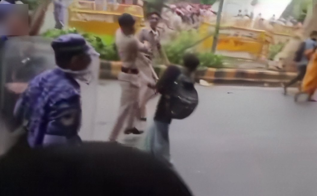 An officer mid-swing with a lathi at a young protester